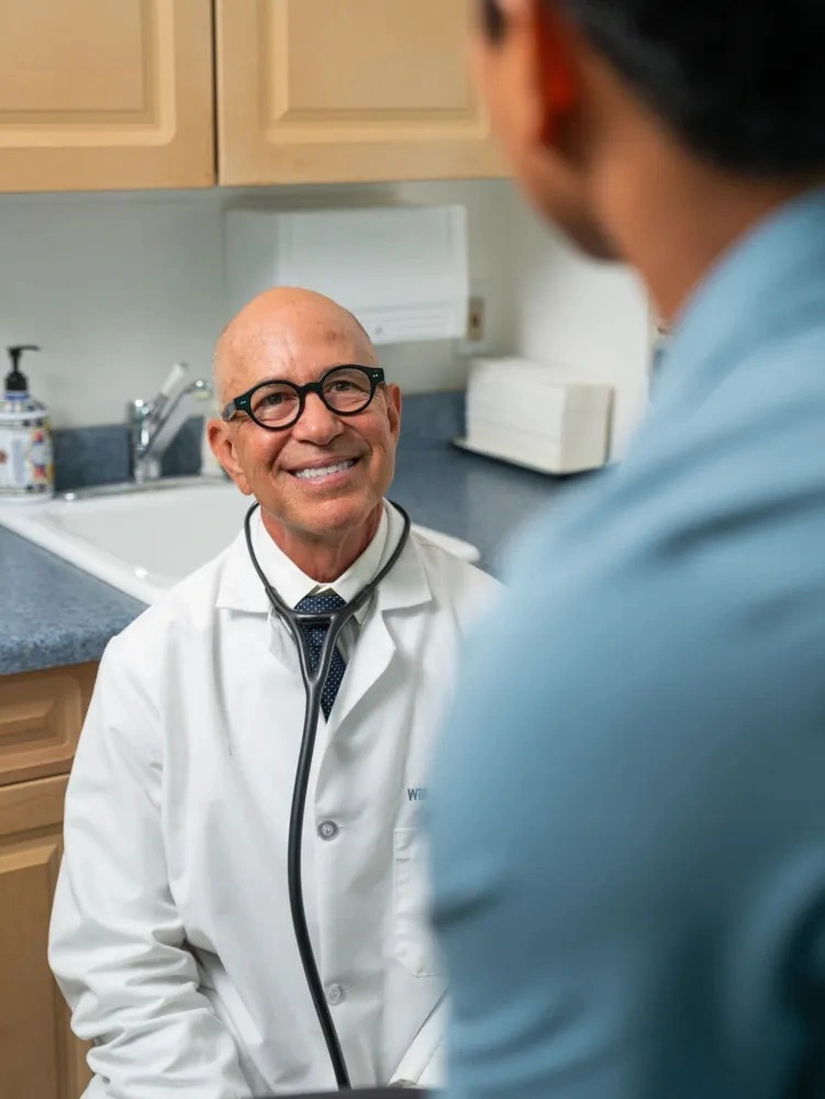 Dr. William Padilla in a white lab coat performing a medical examination with a stethoscope.