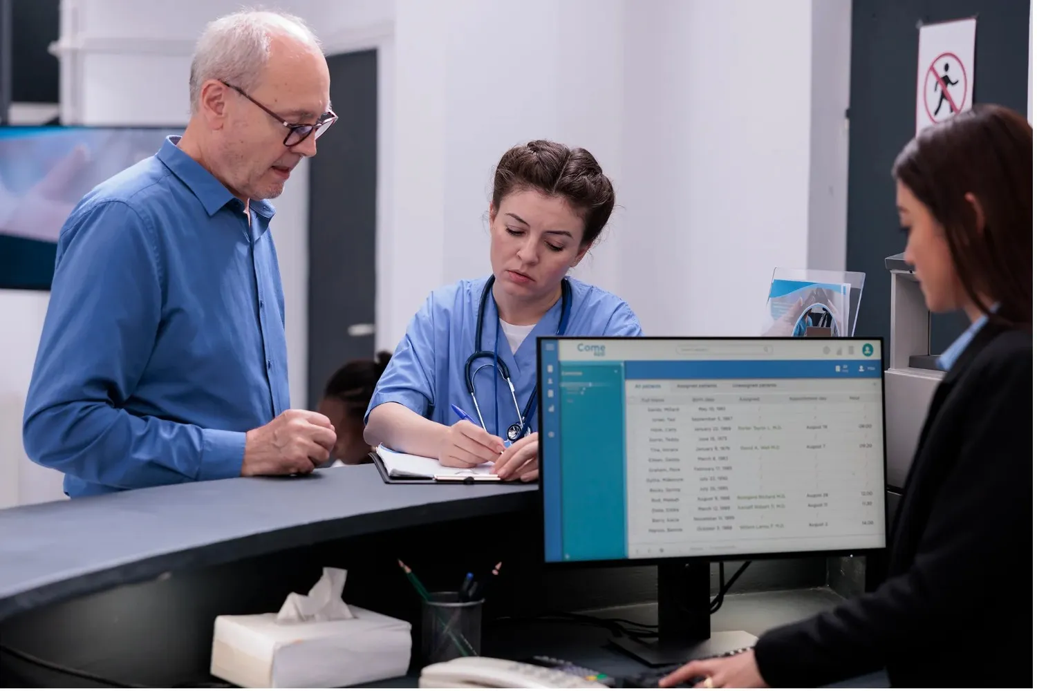 A senior patient speaks with a female nurse in blue scrubs at a clinic reception desk